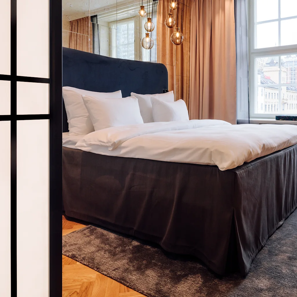 Et hyggeligt hotelværelse med en stor seng, en komfortabel lænestol og et skrivebord ved et lyst vindue.