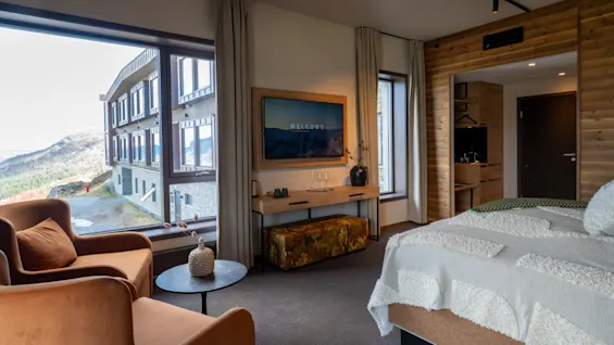 Ett mysigt hotellrum med ett stort fönster som erbjuder utsikt över en byggnad och berg. En TV-skärm visar "WELCOME".