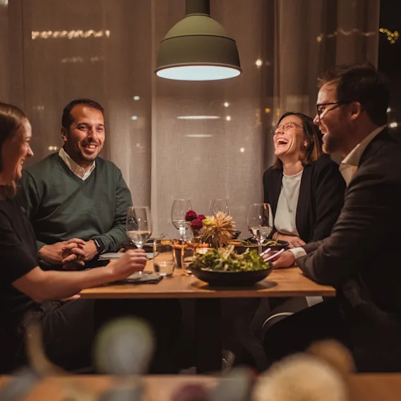 Fire venner som ler og prater rundt et middagsbord i et koselig og varmt opplyst restaurantlokale.