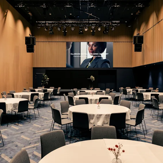 Kokoushuone Clarion Hotel Helsinki Airportissa pyöreillä pöydillä ja tuoleilla, valmiina tapahtumaan. Näyttö näyttää 'Premium jopa 1050'.
