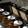 Round marble tables, paired with brown upholstered chairs, line a dark cushioned bench in a sunlit, elegant interior space featuring decorative metal railings and a stone floor.