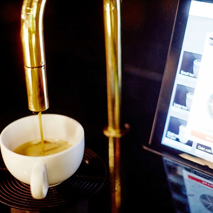 Kaffekop fyldes med espresso fra en gylden dispenser, omgivet af en dæmpet belyst miljø. En digital skærm viser drikkevaremuligheder. Tekst: "Cold milk".