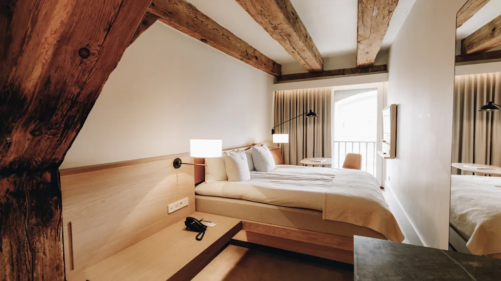 Ett mysigt hotellrum med en bekväm säng, synliga träbjälkar och en ljus balkongutsikt, för en avkopplande vistelse.