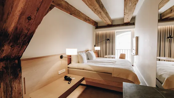 Ett mysigt hotellrum med en bekväm säng, synliga träbjälkar och en ljus balkongutsikt, för en avkopplande vistelse.