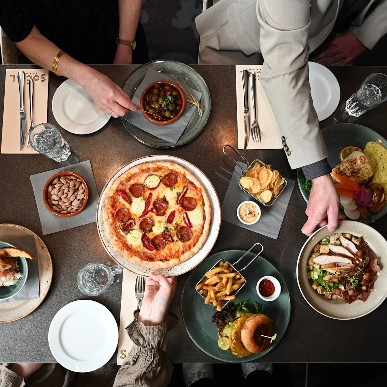 Et bord viser et udvalg af retter, herunder pizza, en burger med pommes frites og en farverig salatskål. To personer rækker efter oliven og justerer tallerkner. "Sorell" er trykt på servietter.