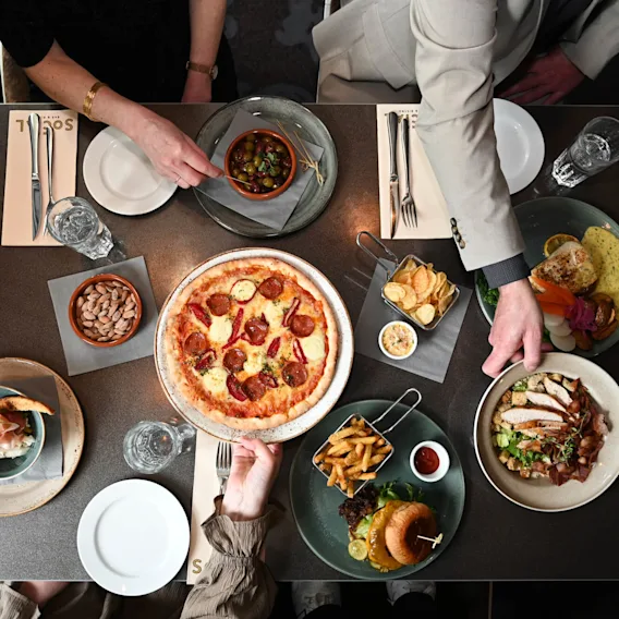 Ett bord visar en mängd olika rätter, inklusive pizza, en hamburgare med pommes frites och en färgglad salladsskål. Två personer sträcker sig efter oliver och justerar tallrikar. "Sorell" är tryckt på servetter.