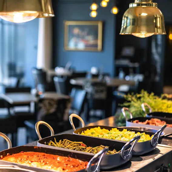 Bufféretter med grøntsager og karry placeret under pendellamper; i baggrunden ses uklare spiseborde og stole i en restaurantomgivelse.