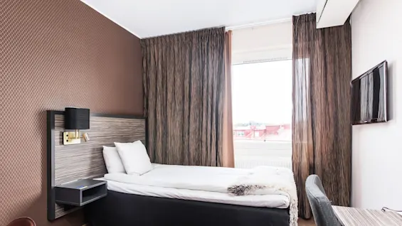 En snyggt bäddad säng i ett rum med mönstrad tapet, gardiner och en platt-TV på väggen, vilket antyder en hotellinteriör.