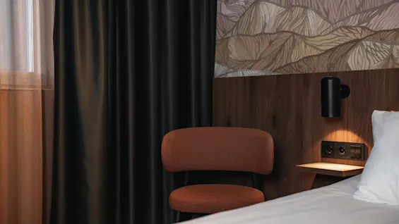 Et moderne hotellrom med en komfortabel seng, en stilig stol, mørke gardiner og en praktisk ladestasjon ved sengen.