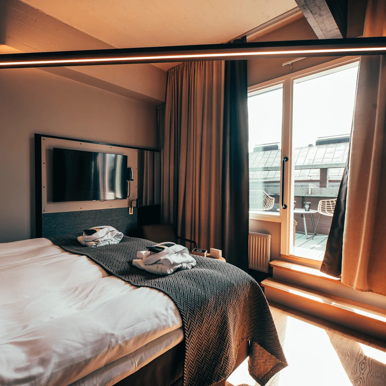 En ryddig seng med et teksturert teppe ligger i et koselig hotellrom. I nærheten er en flatskjerm-TV montert på veggen, og en solfylt balkong er synlig gjennom glassdører.