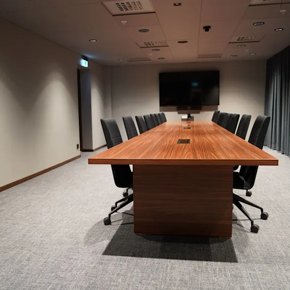 Ett långt konferensbord i trä omgivet av svarta stolar, står i ett modernt mötesrum med grå matta, en stor skärm på väggen och gardiner som täcker ena sidan.
