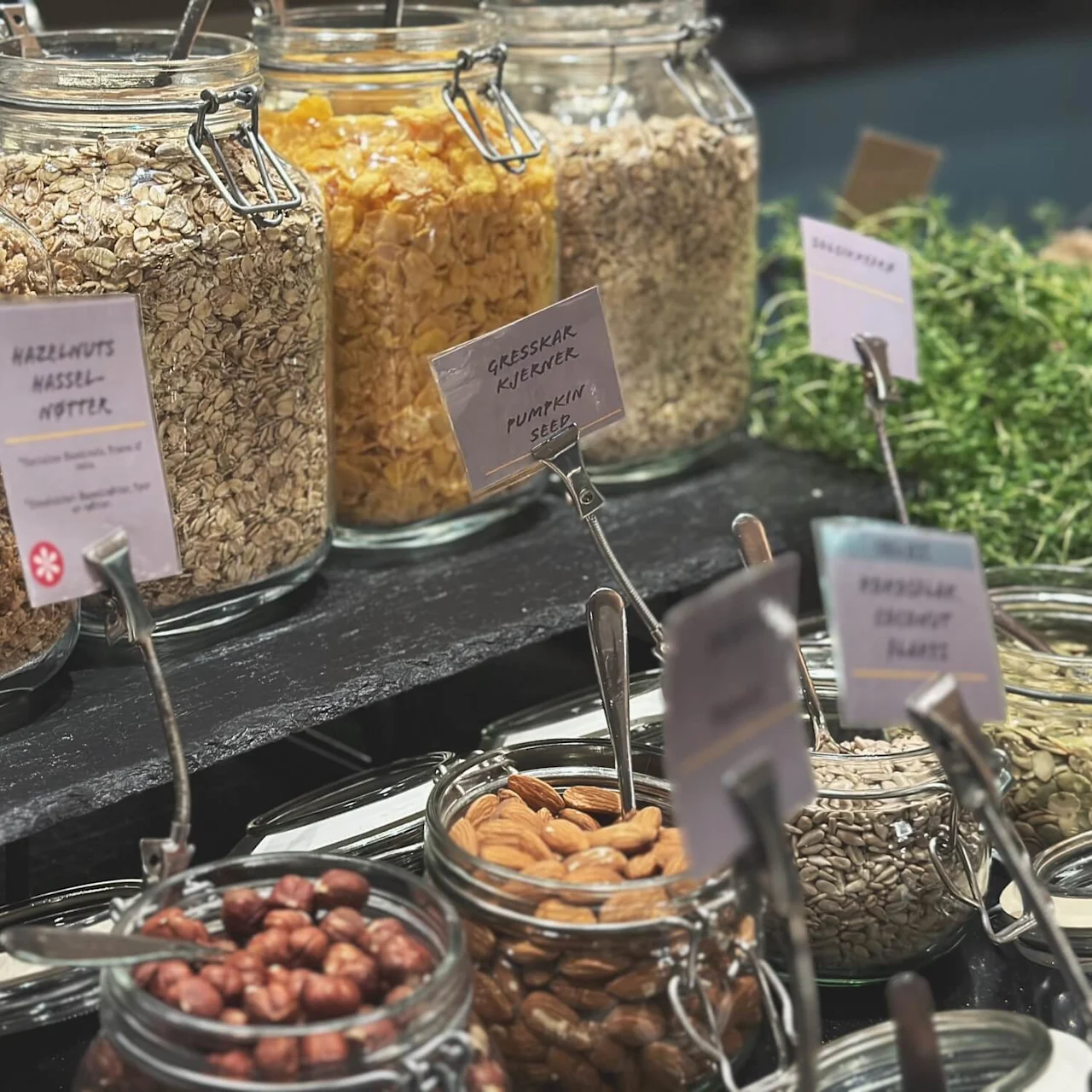 Jars filled with different cereals, nuts, and seeds are displayed on a counter. Labels include "Hazelnuts", "Gresskar Kjerner Pumpkin Seed". Spoons are placed inside the jars for serving.
