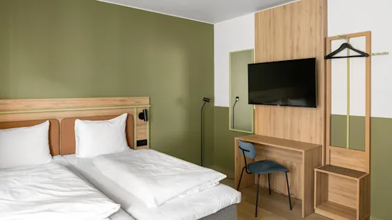 En prydlig hotellrum har en dubbelsäng med vita sängkläder, ett skrivbord i trä, en vägghängd TV och ett grönt och vitt färgschema. En stående lampa och stol kompletterar rummet.