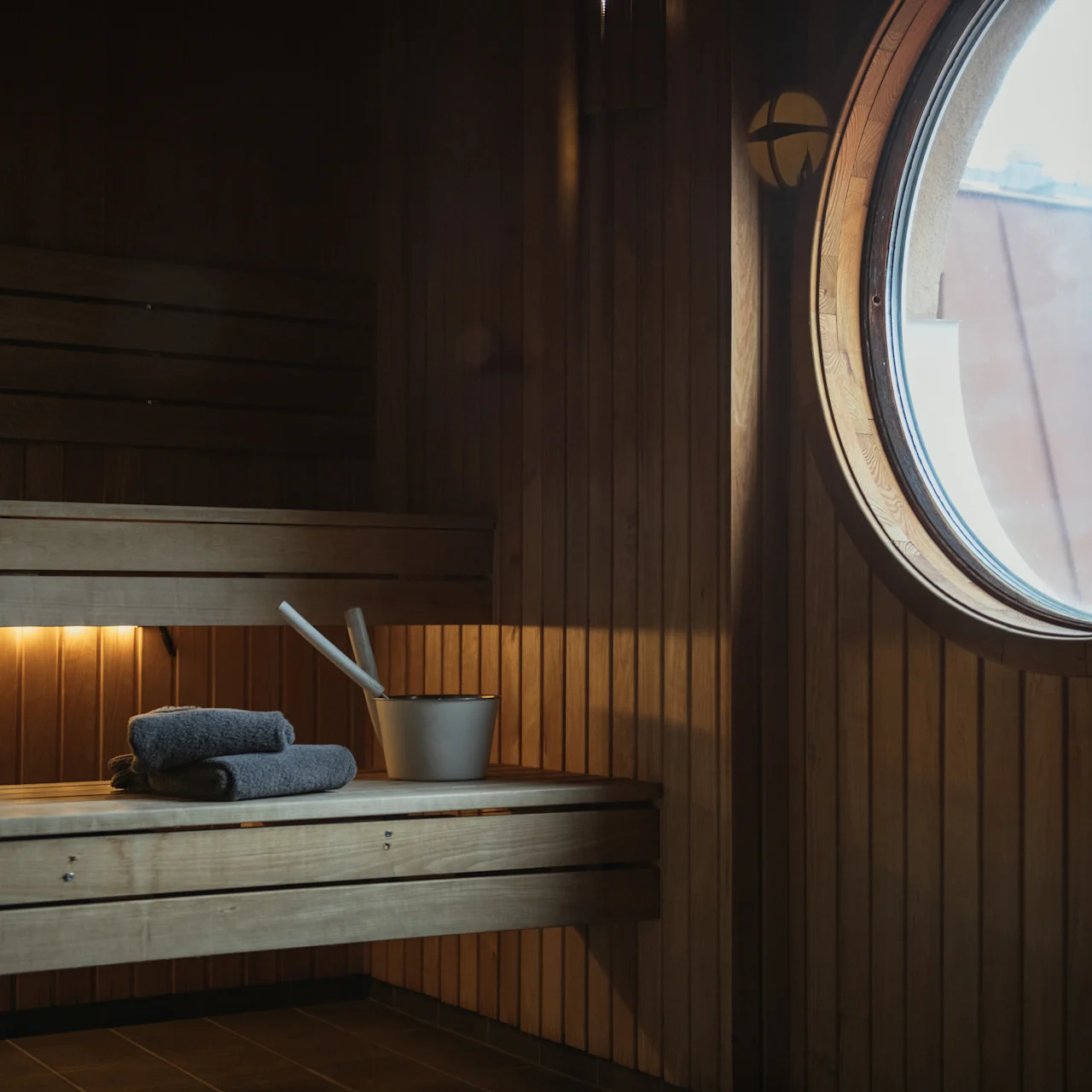 A wooden sauna bench holds two folded towels and a metal bucket with a ladle. Soft lighting beneath the bench enhances the warm atmosphere. A round window showcases an exterior view.