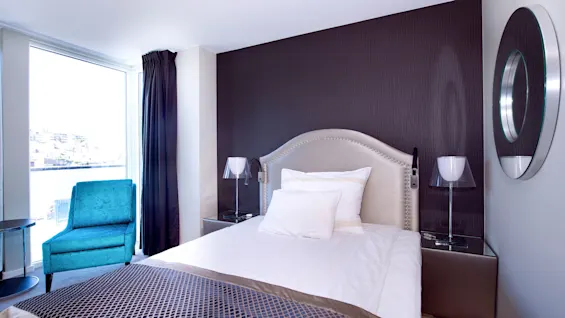 A neatly made bed with a blue armchair, under bright lighting, inside a modern bedroom with a large window.