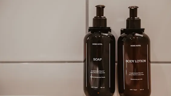 Två mörka flaskor med tvål och body lotion i ett badrum. Etiketterna lyder: SOAP, Gently cleanses your hands, BODY LOTION, Moisturizes your skin.