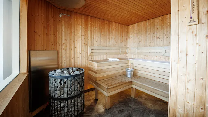 Et trebekledd badstuerom har en hjørnebenk med et håndkle og bøtte, en steinovn som avgir varme, og et veggtermometer med teksten "SAUNA", noe som skaper en koselig atmosfære.