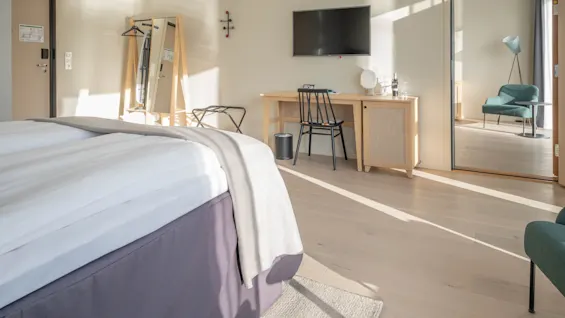 Et lyst og moderne hotelværelse med en komfortabel seng, et skrivebordsområde og en åben døråbning til en lounge.