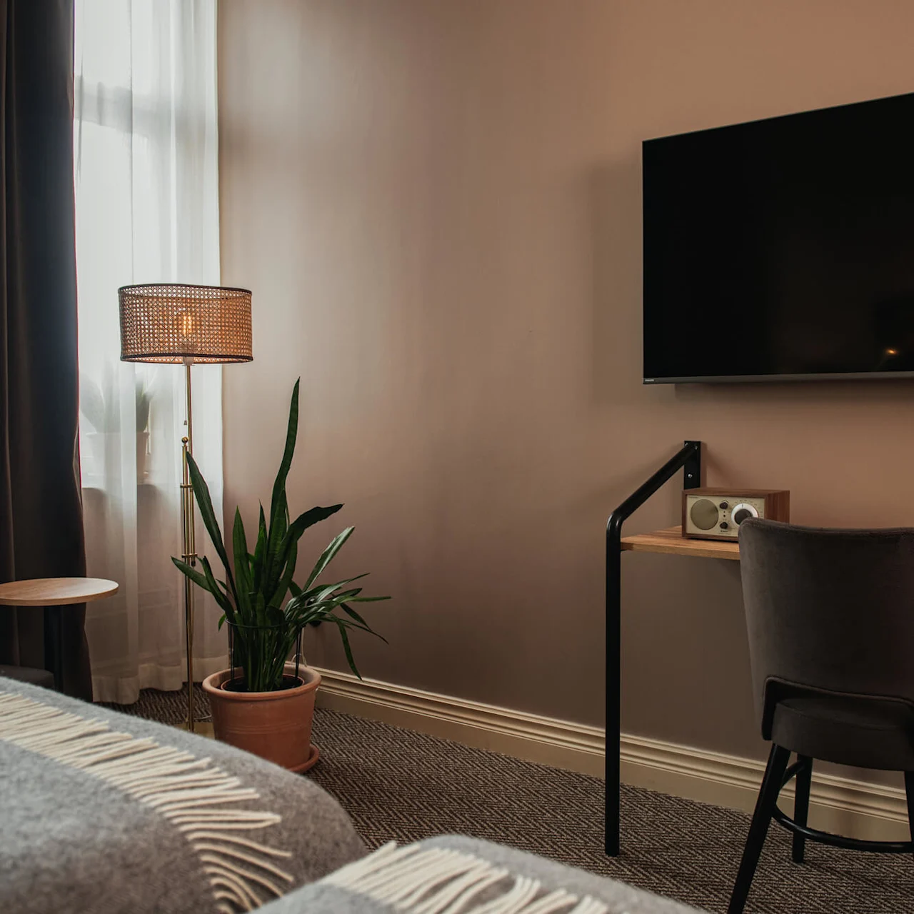 TV on wall above desk with chair, next to a potted plant, beside a floor lamp and a window with sheer curtains in a cozy, modern room.