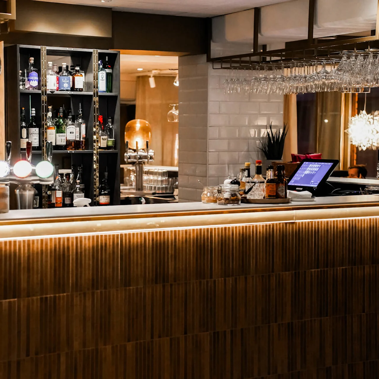 Glas hänger upp och ner ovanför en modern bar fylld med spritflaskor. En digital kassa står på disken, omgiven av ambient belysning i ett varmt upplyst utrymme.