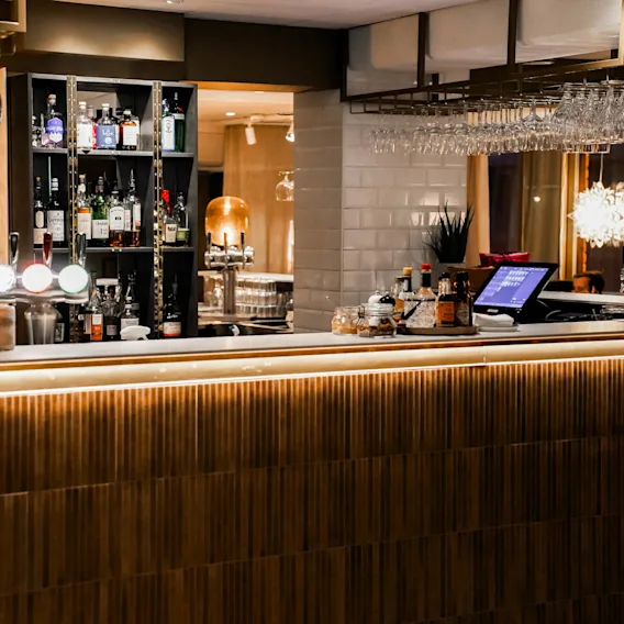 Glas hänger upp och ner ovanför en modern bar fylld med spritflaskor. En digital kassa står på disken, omgiven av ambient belysning i ett varmt upplyst utrymme.