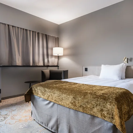 Säng står i ett mysigt, modernt hotellrum, dekorerad med vita lakan och ett guldfärgat överkast. Bredvid lyser en lampa upp de neutrala väggarna och gardinerna.