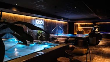 Lyxigt inomhusspa med upplysta pooler, omgivna av stämningsbelysning och modern inredning, inklusive växter och sittplatser. Neonskylt med tre cirklar på väggen ger subtil belysning.