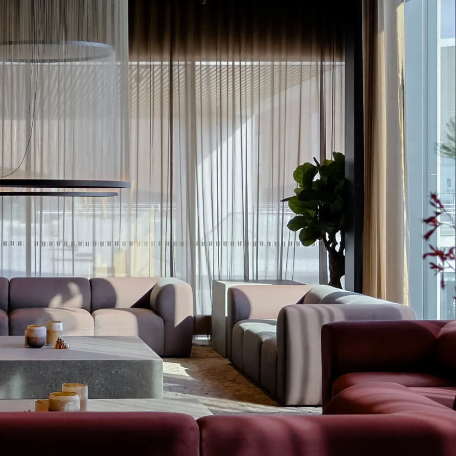 Large gray sectional sofas form a square around a rectangular stone coffee table with candles, in a modern living room with sheer curtains and a hanging circular light fixture.