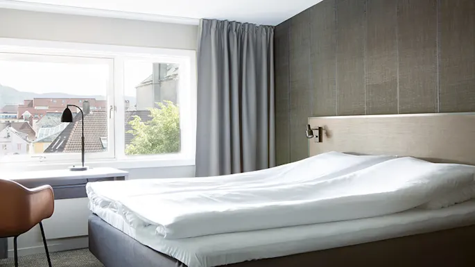 Bed with white linens sits against a textured wall; beside it, a desk and chair face a window overlooking rooftops and greenery.