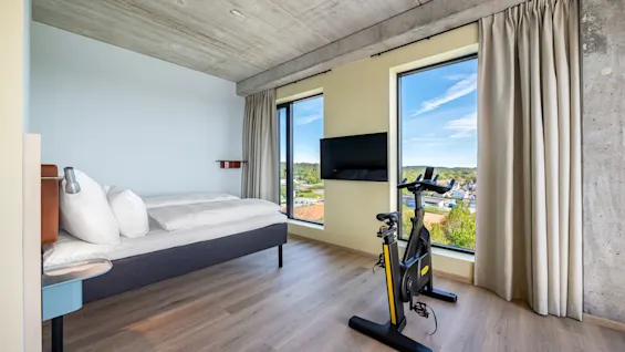 A modern hotel room with a bed, a television, and an exercise bike, offering a comfortable stay with fitness options.