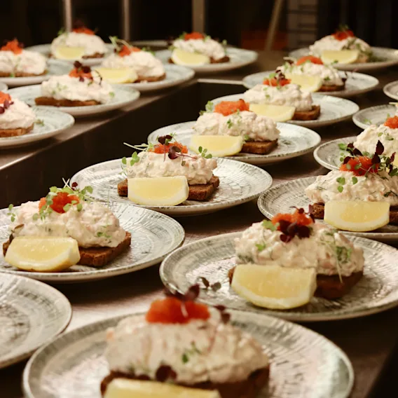 Öppna smörgåsar med krämig skaldjurssallad och citronklyftor vilar på keramiska tallrikar, garnerade med örter, arrangerade i rader på ett restaurangkök.