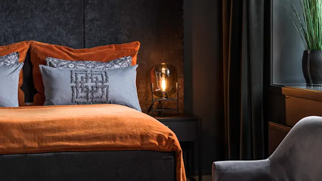 En pent oppredd seng med oransje sengetøy og dekorative puter står ved siden av en tent bordlampe. En grå stol er i nærheten, og en plante pryder vinduet.