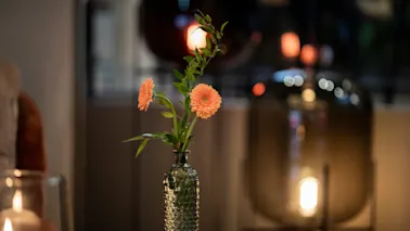 En koselig kveldsscene på Home Hotel Grand Bodø med en vase med oransje blomster på et bord og varme, uskarpe lys i bakgrunnen.