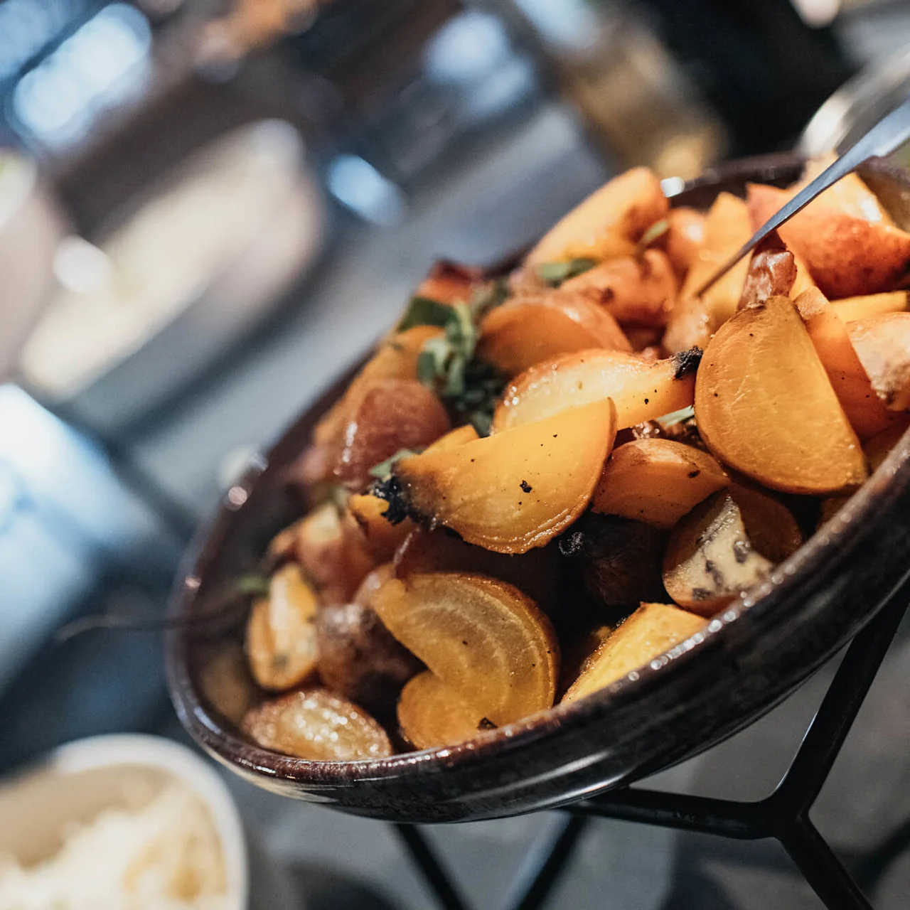 Stekte poteter og urter i en bolle, fokusert i forgrunnen, med en uskarp restaurantinnstilling i bakgrunnen.