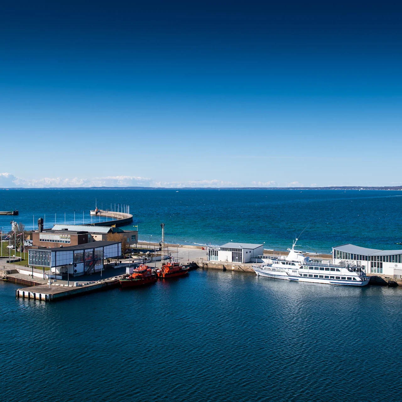 En liten havn med et fyrtårn og bygninger ligger ved det blå havet under en klar himmel. Flere båter og en pir strekker seg ut i vannet.