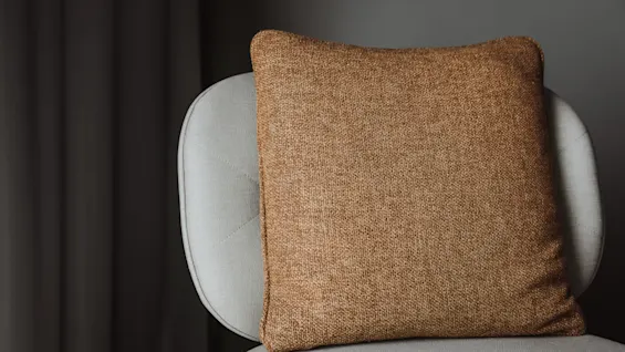 Brun kudde på en ljusgrå stol, placerad mot en mörk bakgrund med gardiner, vilket skapar en mysig och minimalistisk atmosfär.