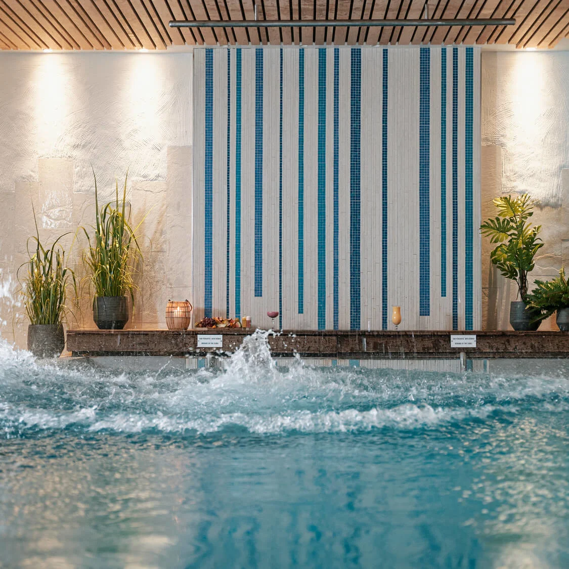 Poolvand sprøjter energisk mod en dekoreret indendørs spa-væg med blå fliser og planter. Varm belysning og træloft giver en afslappende atmosfære.