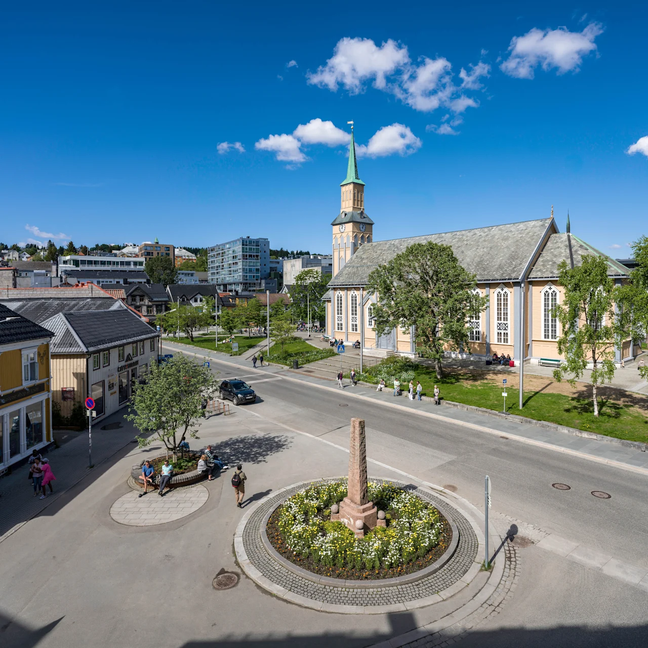 Church stands near road, surrounded by trees and grass. People walk and sit nearby. Buildings, including a souvenir shop, line the street under a clear blue sky.
