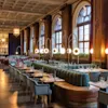 Bord och fåtöljer arrangerade i en elegant restaurang, upplysta av stilfulla hänglampor, med stora valvformade fönster längs väggen, vilket ger en sofistikerad och stämningsfull matmiljö.