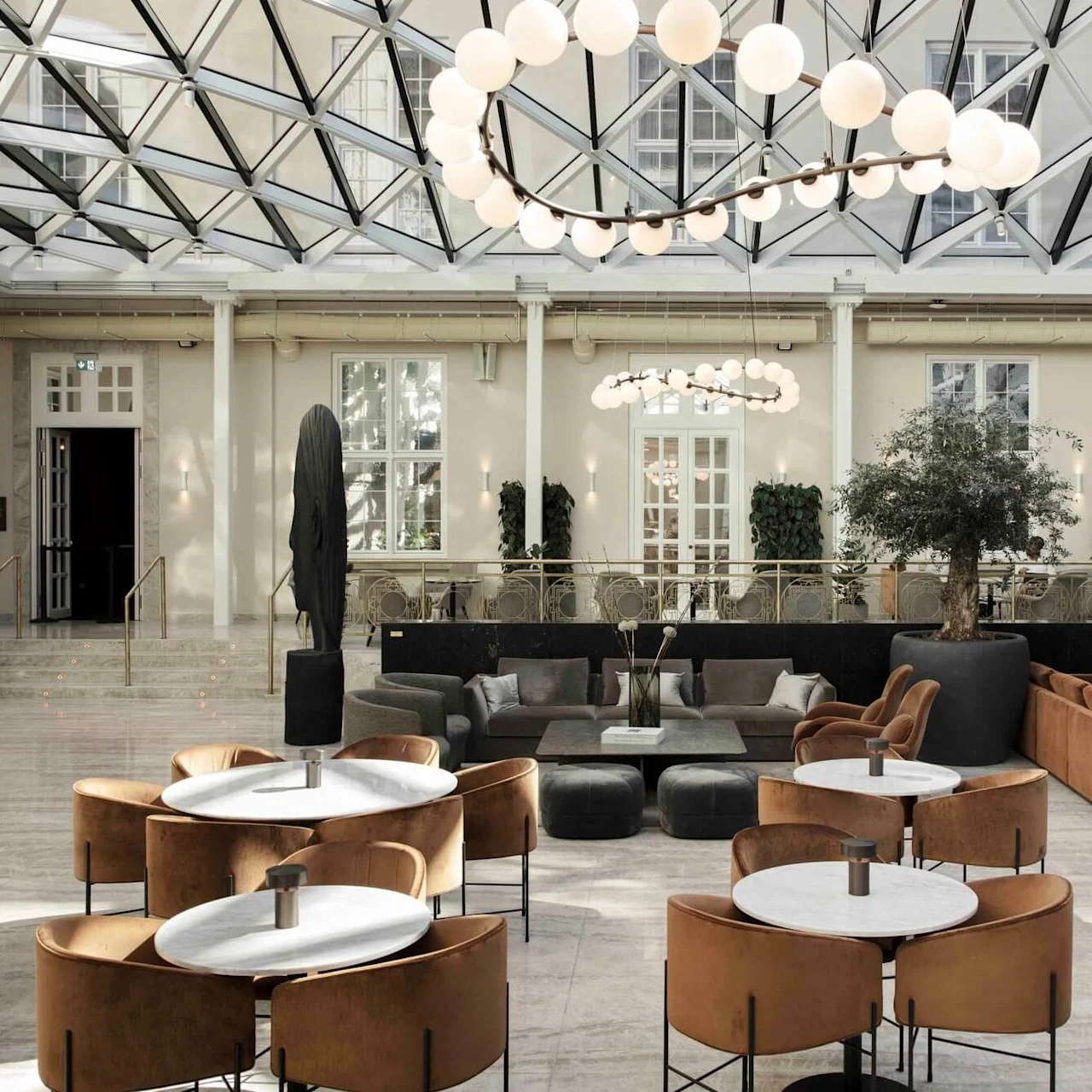 Tables with round marble tops are surrounded by leather chairs, situated in a spacious indoor lounge under a geometric glass ceiling, with cozy sofas, plants, and a sculpture nearby.