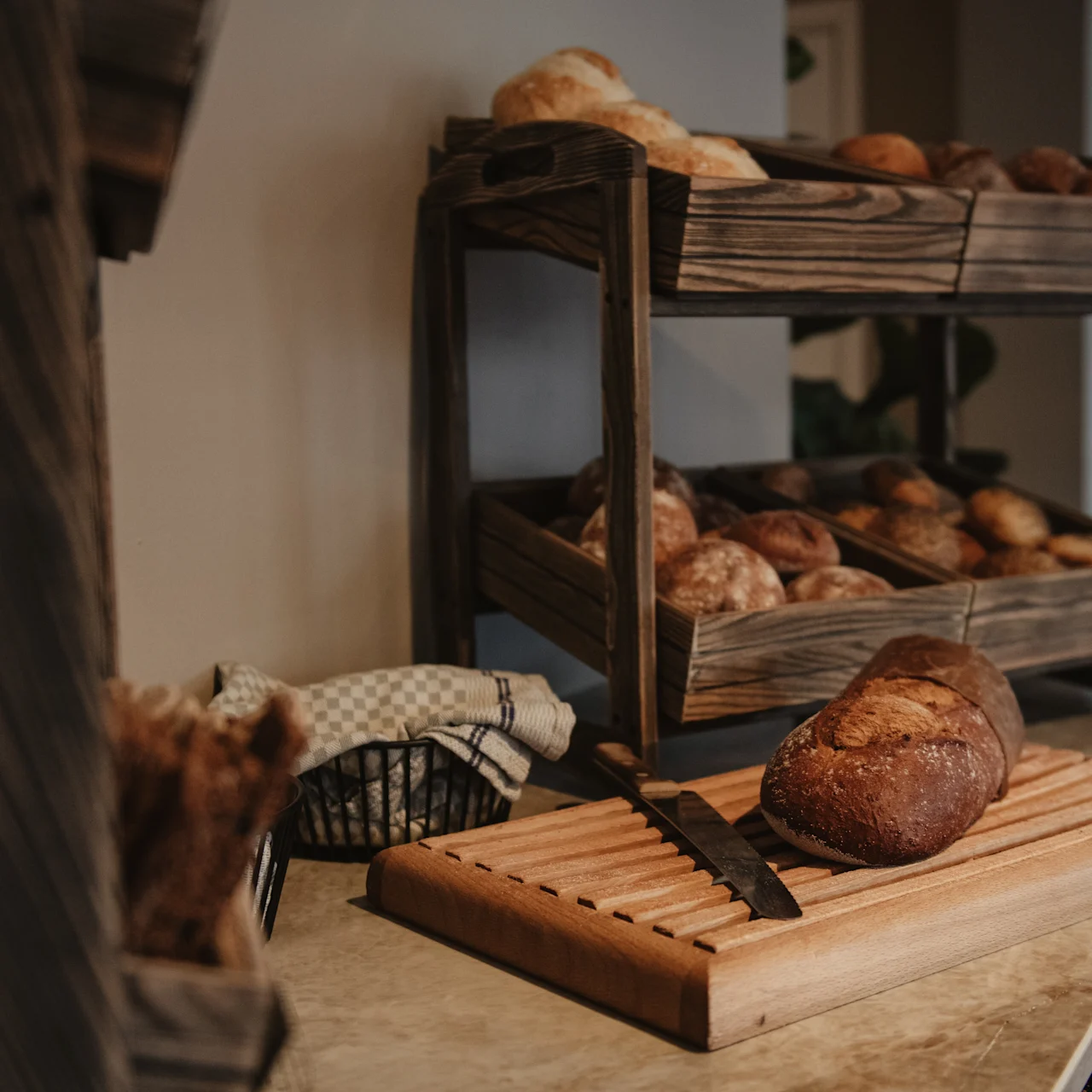 Bread sits on a wooden cutting board with a knife, surrounded by stacked wooden racks filled with various loaves, in a rustic kitchen setting.