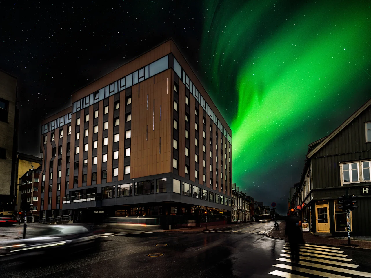 Yökuva rakennuksesta nimeltä THE EDGE eloisan revontulen alla, katu valojuovineen ja kävelevä henkilö. 