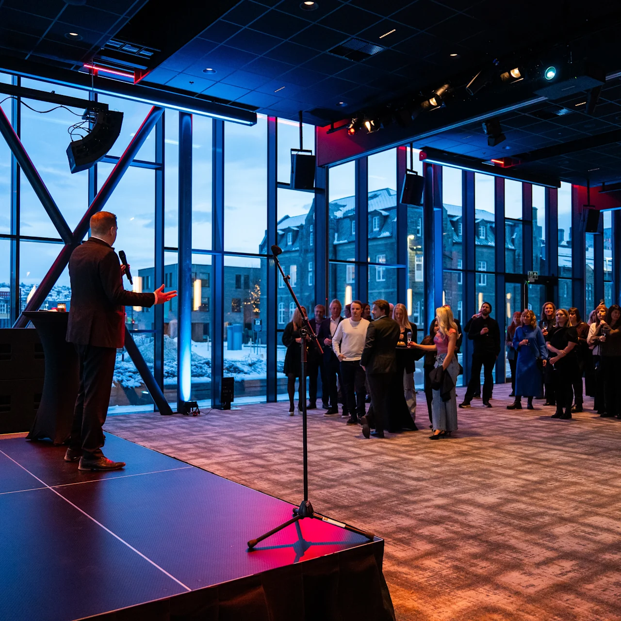 A person speaks on a stage, addressing a crowd in a large, modern room with floor-to-ceiling windows overlooking a snowy urban landscape.