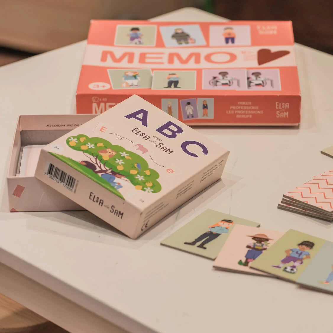 Boxes of educational cards lie on a table, with scattered illustrated character cards. Text: "MEMO," "Elsa och Sam," "YRKEN PROFESSIONS LES PROFESSIONS BERUFE," "ABC med ELSA och SAM."