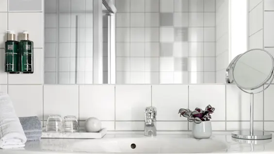 A bathroom sink with a silver faucet is surrounded by a mirror, two bottles of hand wash, neatly folded towels, glasses, a marble tray, a plant, and a round makeup mirror.