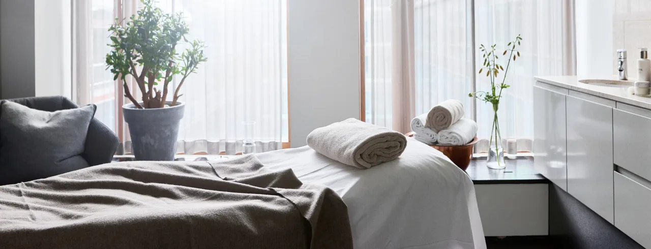 A neatly made bed with towels, next to a window letting in daylight, in a tidy room.