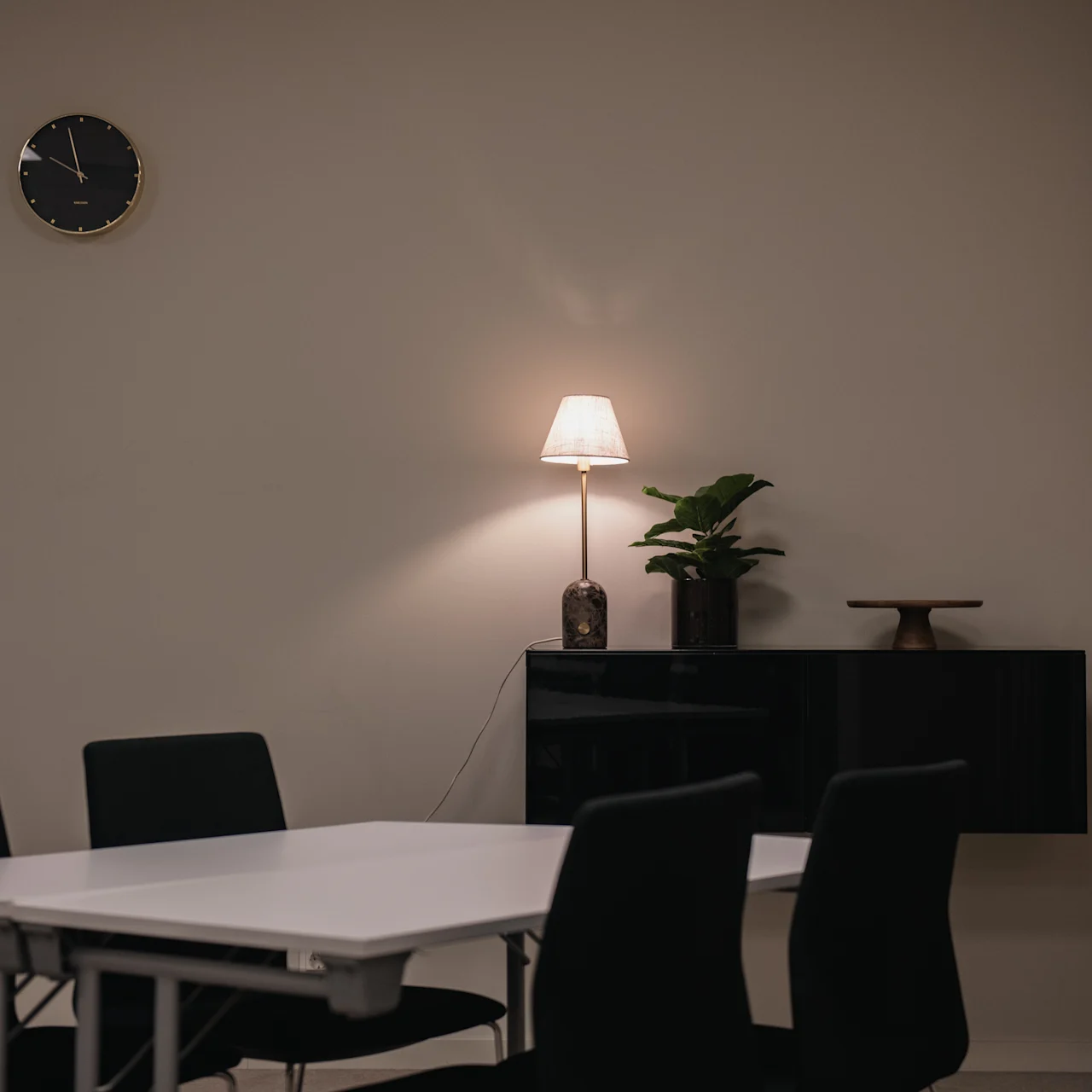 A white table surrounded by black chairs is situated in a minimalist room. A wall clock and a lamp with a plant and decor are on a shelf nearby.