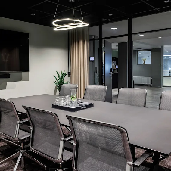 Et moderne møterom med et langt grått konferansebord, grå nettingsstoler, en sirkelformet taklampe og stor skjerm.