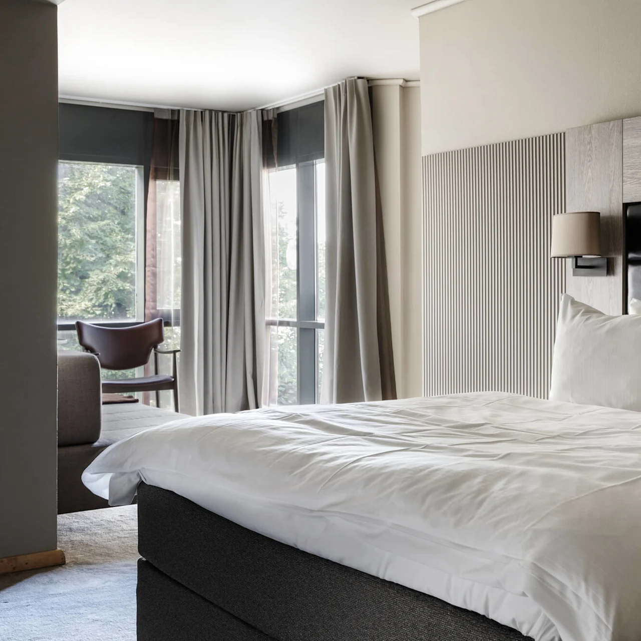 A neatly made bed with white linens and pillows, positioned in a modern hotel room with large windows, draped gray curtains, and a chair in the corner near a sofa.