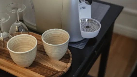To keramikkopper står på en trækbakke ved siden af en espressomaskine, placeret på et mørkt bord med to omvendte vinglas i nærheden.